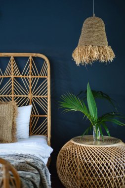 Etnik dekorlu otantik yatak odasının dikey fotoğrafı, komodinin üzerinde lamba, yatak ve doğal yeşil bitki kompozisyonu. Rahat evdeki oda kavramı