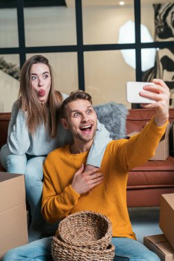 Genç ve komik bir ailenin dikey fotoğrafı yeni bir daireye taşınıyor, modern akıllı telefonda fotoğraf çekiyor, eğleniyor, taşınmayı kutluyor, kutuların yanındaki koltukta oturuyor.