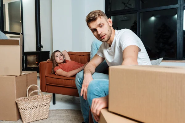 Real estate concept. Tired and exhausted young adult newlyweds move in new house together, sitting near cardboard boxes, resting in living room with modern couch