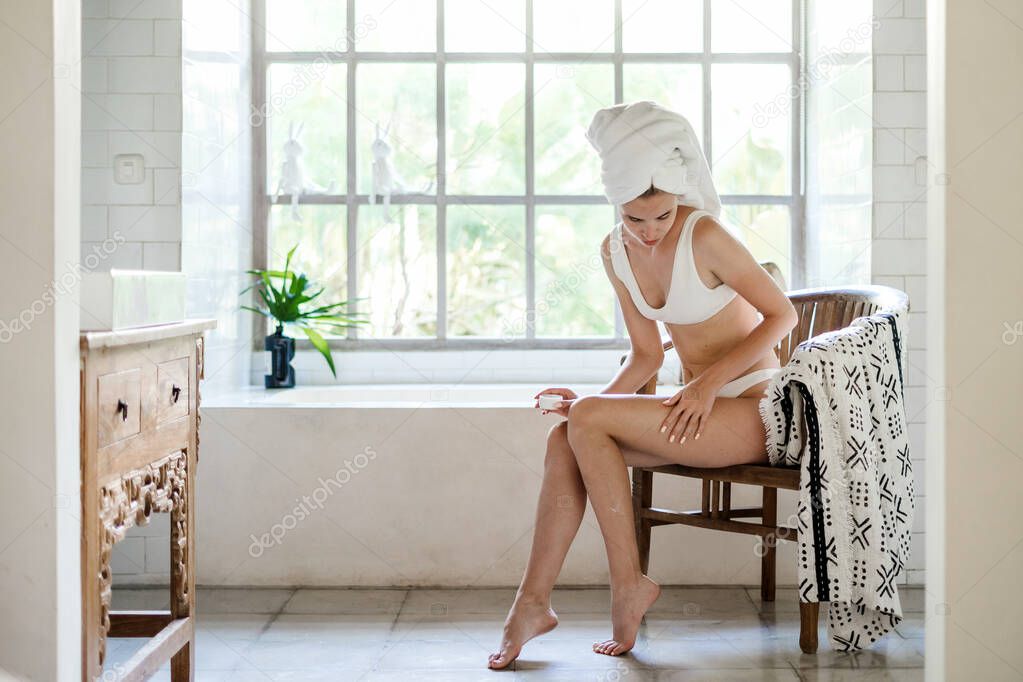 Young adult girl with towel on head sitting on chair in white bathroom, applying soft natural cream on legs, making skin and body care procedure