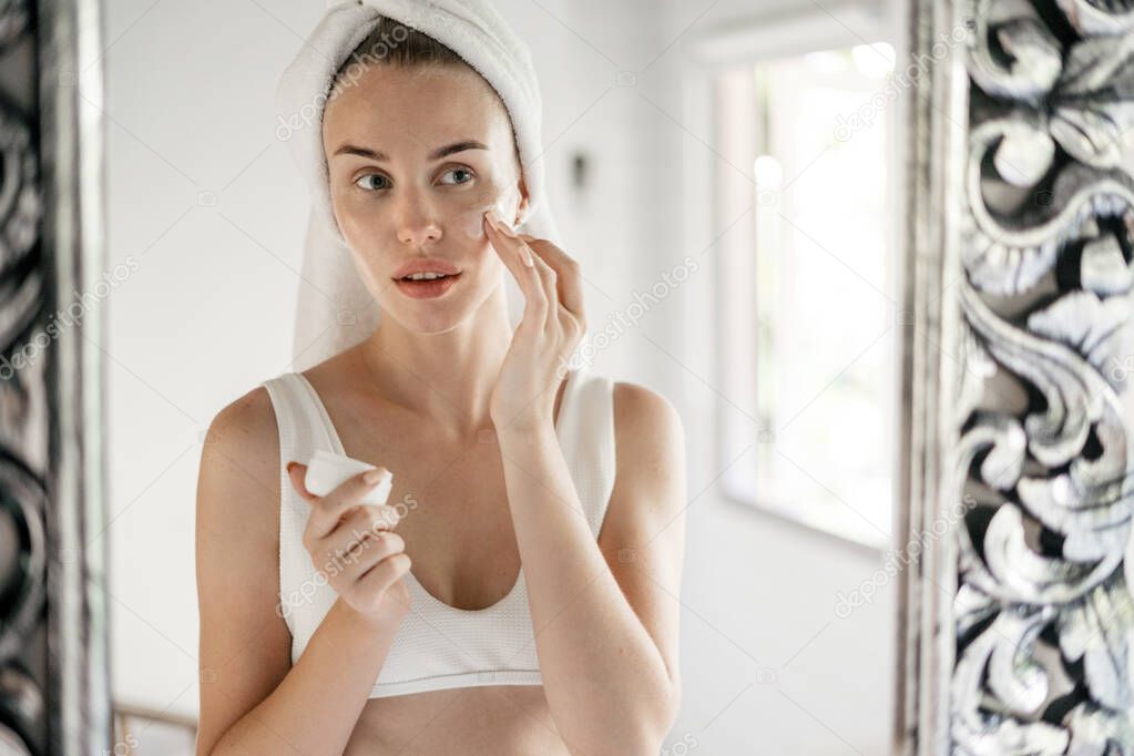 Portrait view of young adult girl applying natural face cream on soft clear skin, looking at mirror, standing in white bathroom with towel on head