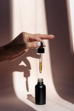Kırpılmış ve dikey olarak genç bir kadının cam şişenin üzerinde doğal yağla pipeti tutarken görüntüsü, fotokopi alanı olan pastel pembe arka plana serum dökerken görüntüsü.