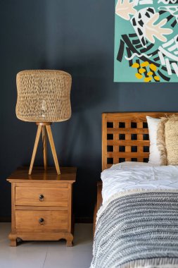 Etnik dekorlu rahat bir ev. Klasik yatak odasının dikey fotoğrafı ahşap komodinin üzerinde lamba, rahat bir yatak ve duvarda resim.