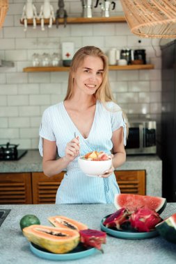 Sağlıklı ve mevsimsel gıda konsepti. Mutfakta dikilen, kahvaltıda meyve salatası yiyen ve gülümseyen genç bir kadının dikey fotoğrafı.