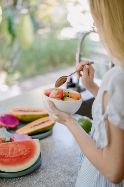 Sağlıklı ve mevsimsel beslenme kavramı. Mutfakta elinde kahvaltı kâsesiyle duran meyve salatası yiyen ve yemeğin tadını çıkaran genç bir kızın dikey ve kırpılmış fotoğrafı.