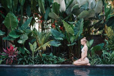 Güzel yetişkin bir kızın yaz gününü yüzme havuzunun yanında, bikini mayosuyla havuz kenarında yeşil bahçe arka planında yaprakları ve doğal yeşilliklerle oturarak geçirmesinin yan görünüşü.