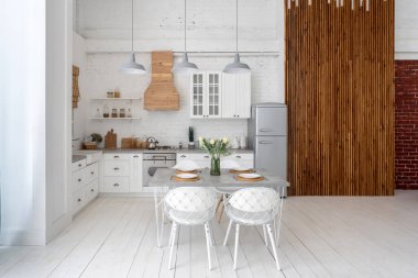 Modern iç mekanı, dolapları, yeni mobilyaları, yemek masası ayarı, gaz ocağı, fırın ve ahşap panel duvarının yanındaki buzdolabı manzaralı beyaz mutfak.