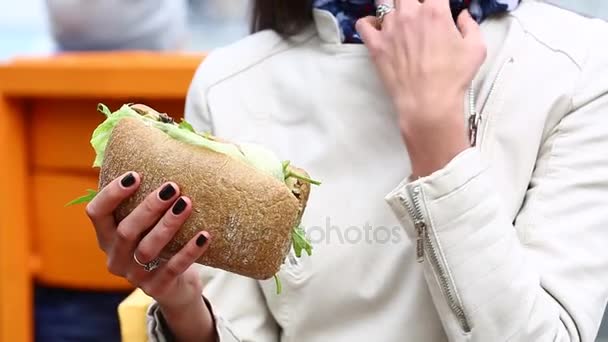 Fiatal nő evés szendvics kávézóban a szabadban. Szép nő evés szendvics kávézóban. Vonzó nő enni egy szendvicset, a parkban, zenehallgatás — Stock videók