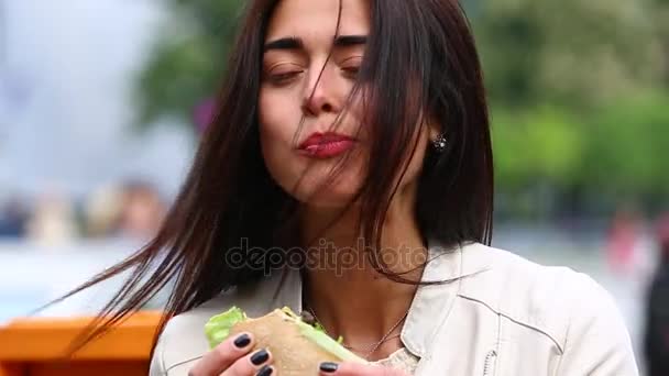 Mujer joven comiendo sándwich en la cafetería al aire libre. Una mujer guapa comiendo sándwich en la cafetería. Atractiva mujer comiendo un sándwich en el parque, escuchando música — Vídeo de stock
