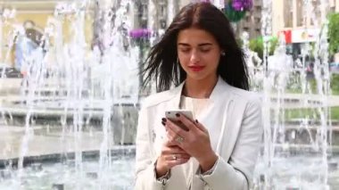 Smartphone kullanarak gülen kız portresi. Cep telefonu çeşme arka plan üzerinde olan genç ve güzel kadın. Şehir Parkı, atış Steadicam Smartphone kullanan oldukça mutlu kadın.