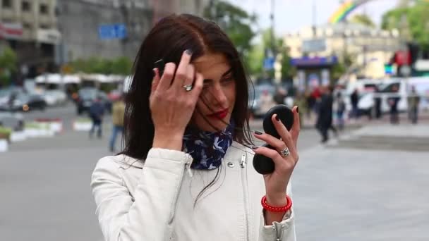 Een mooi meisje in een witte regenjas schildert haar lippen met lippenstift. Aantrekkelijke jonge dame terwijl het zoeken op de spiegel maken haar lippen rood en sexy door een nieuwe glamour lippenstift op straat. — Stockvideo