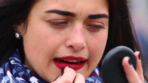 Een mooi meisje in een witte regenjas schildert haar lippen met lippenstift. Aantrekkelijke jonge dame terwijl het zoeken op de spiegel maken haar lippen rood en sexy door een nieuwe glamour lippenstift op straat. — Stockvideo