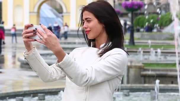 Portret van meisje lachen met behulp van de smartphone. Mooie jonge vrouw met haar mobiele telefoon op de achtergrond van de fontein. Vrij gelukkig vrouw met behulp van smartphone in stadspark, Steadicam schot. — Stockvideo