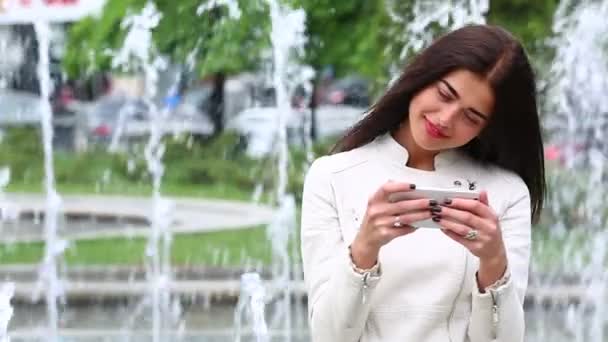 Portret dziewczyny, śmiejąc się, za pomocą smartfonu. Dość młoda kobieta z jej telefonu komórkowego na tle fontanny. Zadowolony, Kobieta za pomocą smartfona w parku miejskim, Steadicam strzał. — Wideo stockowe