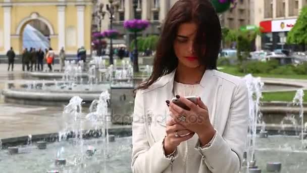 Porträtt av flicka skratta med smartphone. Ganska ung kvinna med sin mobiltelefon på fontänen bakgrund. Ganska nöjd kvinna med smartphone i stadsparken, Steadicam sköt. — Stockvideo