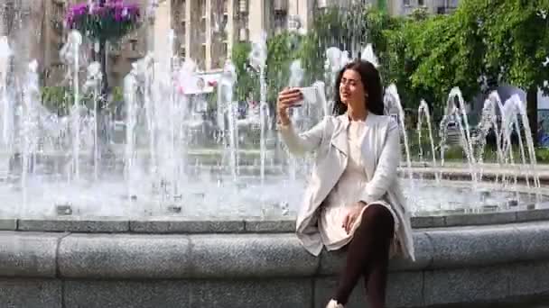 Красива молода жінка зі своїм мобільним телефоном на фоні фонтану . — стокове відео