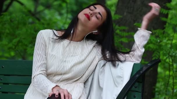 Mujer escuchando la música desde un teléfono inteligente con auriculares en un parque . — Vídeos de Stock