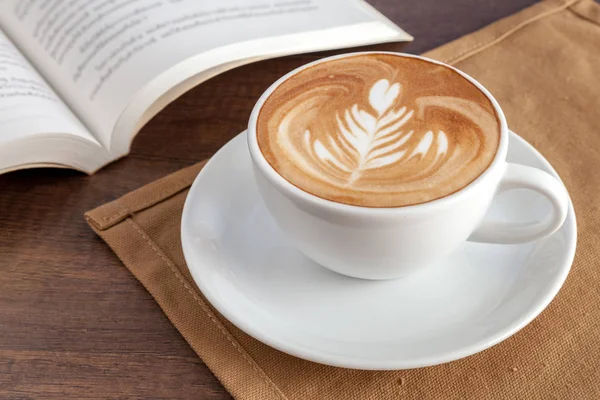 Koffiekopje van rosetta latte art op servet met een boek naast. — Stockfoto