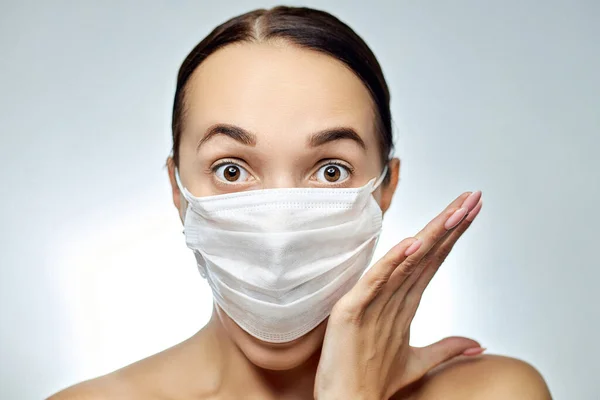 Young brunette girl with a medical white mask on her face with a mask and hands next to her looks surprised ahead — Stock Photo, Image