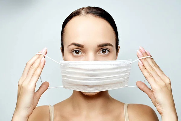 Güzel bir kız gri bir arka plana tıbbi maske takar ve kendine güvenen düzgün bir saç stili takar.