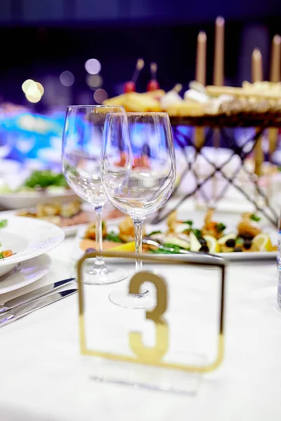 На праздничном столе с белой скатертью два кристально чистых стакана для напитков и металлическая пластина с номером три — стоковое фото