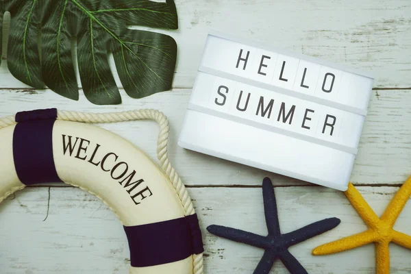Hello Summer text in light box with green leave, starfish and lifebuoy decoration on wooden background