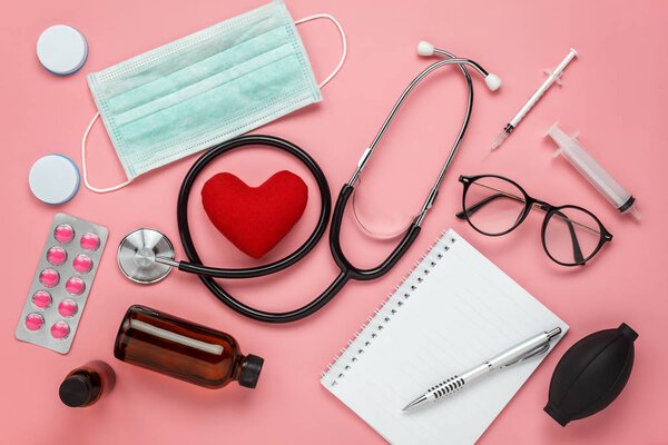Плоское аэрографическое изображение аксессуаров здравоохранения и медицинского фона, скрытое красным сердцем и стетоскопом с оборудованием на розовой бумаге.
.