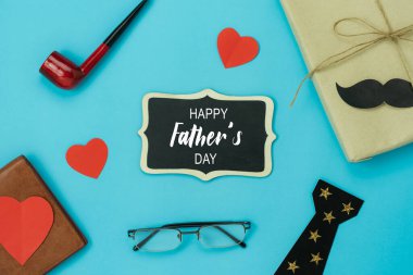 Tablonun tepesinde Mutlu Babalar Günü tatili konsepti. Babam için aksesuar hediye kutusu. Mevsim nesneleri, cüzdan, tütün piposu ve mavi kağıt üzerinde tebrik kartı..