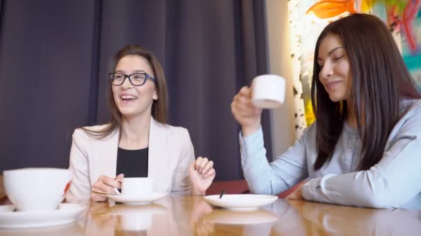zwei Frauen am Tisch hören zu, lächeln und trinken ein Getränk aus weißen Bechern.