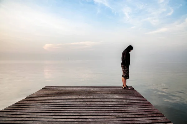Joven deprimido parado en un puente de madera extendido en el mar mirando hacia abajo — Foto de Stock