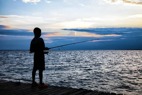 Silhouette of the boy fishing on wooden bridge extended into the sea on sunset. — Stock Photo, Image