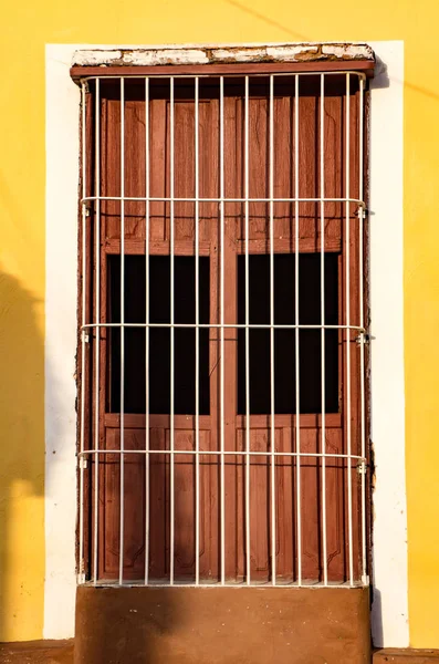 Brown shuttered window behind white metal bars on yellow and whi