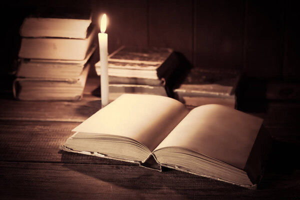 An open book with blank pages lies on a wooden table on the background of other standing books and burning candle.