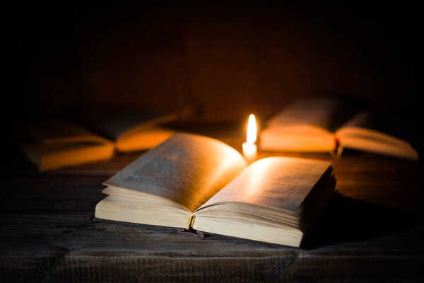 An open book with blank pages lies on a wooden table on the background of other opened books and burning candle.