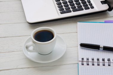 Bir fincan kahve, beyaz masadaki kalem ve dizüstü bilgisayarlı defter.