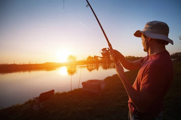 Молодой человек рыбачит на озере на закате наслаждаясь хобби — стоковое фото