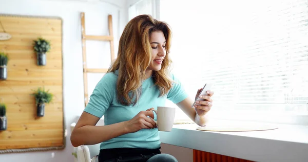 Beautiful woman using cell phone and drinking morning coffee at home