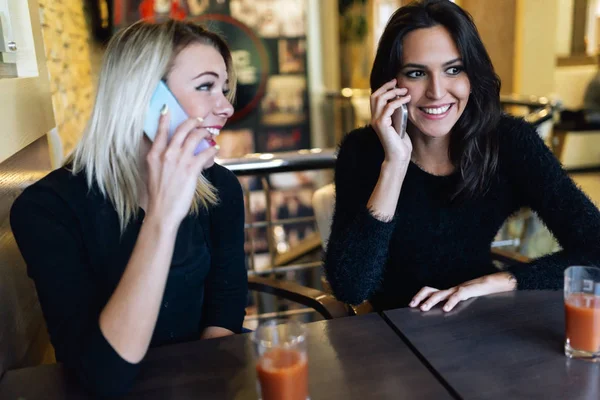 Two beautiful women talking on phone in cafe while being happy