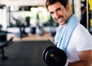 Spor salonunda egzersiz yapan kaslı, formda bir adam. İki kollu halterle egzersiz yapıyor.