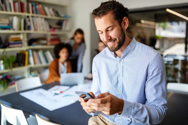 Joven Feliz Mirando Teléfono Móvil Sonriendo Mientras Sus Colegas Trabajan — Foto de Stock