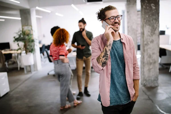 Joven Feliz Mirando Teléfono Móvil Sonriendo Mientras Sus Colegas Trabajan — Foto de Stock