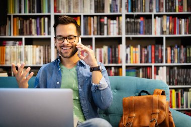 Zevkle çalışıyorum. Cep telefonuyla konuşan ve dizüstü bilgisayarına bakan mutlu genç adam.