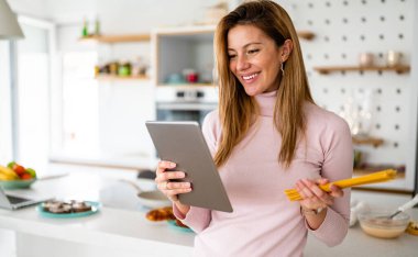 Gülümseyen kadın dijital tablette tarifine bakıyor, mutfakta yemek pişirmeye hazırlanıyor.