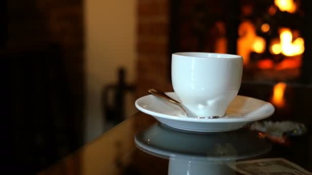 Cup of coffee is on table in cafe or restaurant with fireplace. Glimmers of flame reflect on the table. Romantic setting. Selective focus. Close-up. — Stockvideo