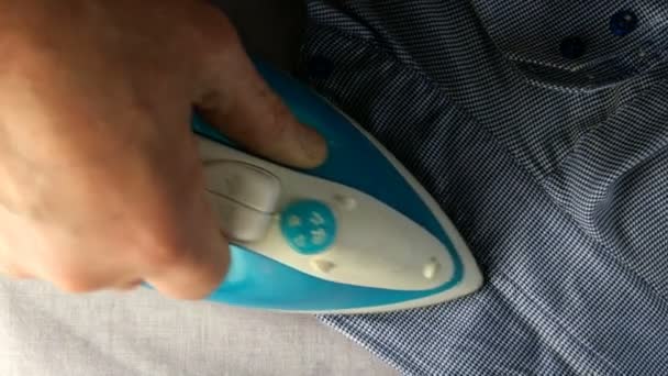 Menselijke hand met elektrisch strijkijzer is strijken blauw shirt, op lichte textiel achtergrond. Bovenaanzicht. Huishoudelijk concept. Close-up. — Stockvideo