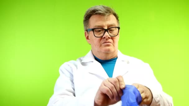 Серьезный старший взрослый врач в белом халате надел защитные резиновые перчатки, которые защищают от инфекции снаружи. Зеленый фон. Хромаки. Крупный план . — стоковое видео