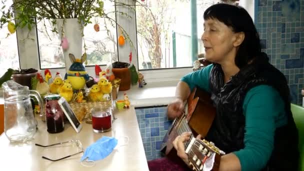 Güzel, son sınıf öğrencisi bir kadın gitar çalıp şarkı söylüyor. Mutfakta pencerenin yanında oturuyor. Ortam planı. — Stok video
