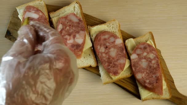 Die menschliche Hand legt eingelegte Gurkenscheiben auf Wurstbrötchen, die auf einem Schneidebrett auf dem Tisch stehen. Lebensmittel von oben nach unten. — Stockvideo