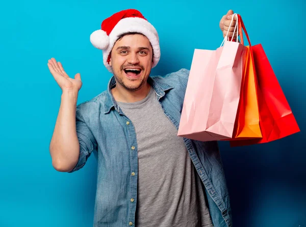 Snygg man i julhatt med shoppingväskor på blå bakgrund — Stockfoto
