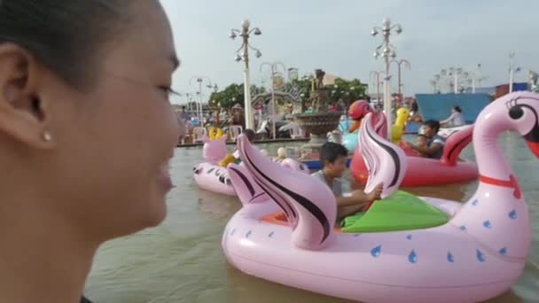 Una madre asiatica guarda i suoi figli multiculturali mentre cavalcano su un cigno gonfiabile in una piscina di un parco divertimenti — Video Stock